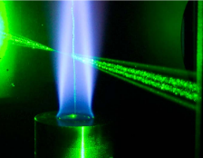Laser-Raman Spektroskopie einer turbulenten Erdgasflamme mit Tropfeneindüsung. Die Erdgasflamme ist durch ihre bläuliche Chemolumineszenz erkennbar. Der Tropfenstrom, erkennbar als grüner vertikaler Strahl, verläuft in der Mitte der Flamme von unten nach oben. Die von links nach rechts verlaufenden Streifen markieren die Anregungs-Laserstrahlen für das laserbasierte Messverfahren.